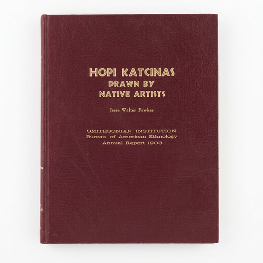 A vintage hardcover book titled 'HOPI KATCINAS DRAWN BY NATIVE ARTISTS' by Jesse Walter-Fewkes,  The book cover is a deep burgundy with gold lettering, placed on a light-colored wooden surface.